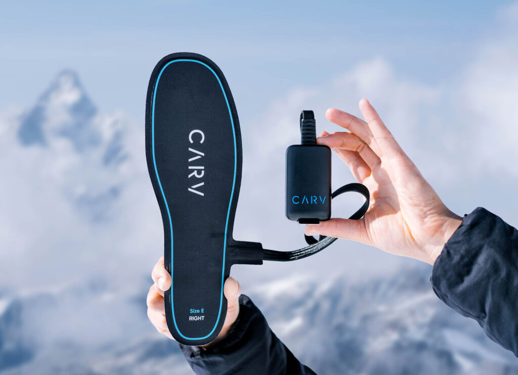 Ski device Carv - a footbed and electric clip device - is held up to the camera, a blurry mountain backdrop