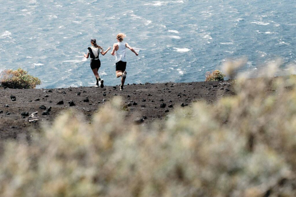 two trail runners, a man and woman, running together away from camera, with a blurred-out bush in the foreground, on scree-like terrain and downhill