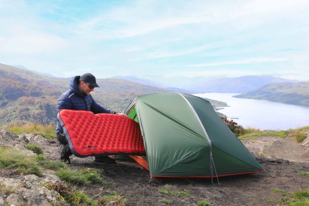 camper pushes red inflated mattress into entrance of a small green tent, pitched on a high plateau overlooking a lake