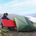 camper pushes red inflated mattress into entrance of a small green tent, pitched on a high plateau overlooking a lake