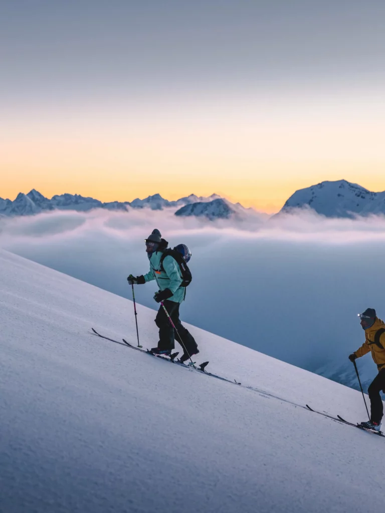 sunrise, high in the mountains, pointed peaks in the distance silhouetted against a sunset or rise, with two ski tourers heading uphill on fresh snow