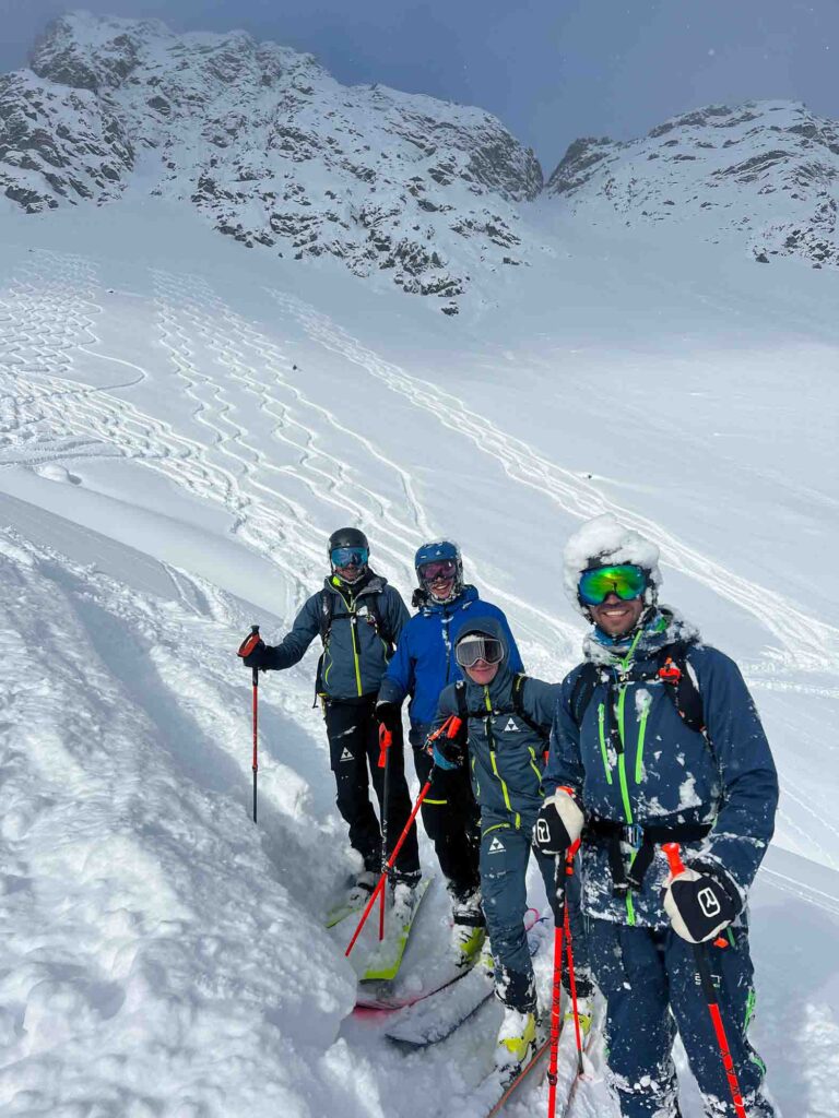 four guys, on the hill, having made fresh tracks side-by-side, the closest to camera wearing a halo of snow on his helmet