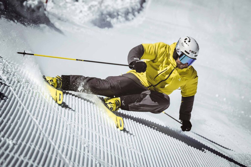 a skier in yellow in a deep carve on fresh groomed corduroy, showing the yellow of the base of his skis