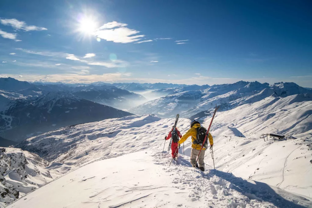two skiers, at the top of a mountain, skis on backs, boot downhill looking over a huge mountain view