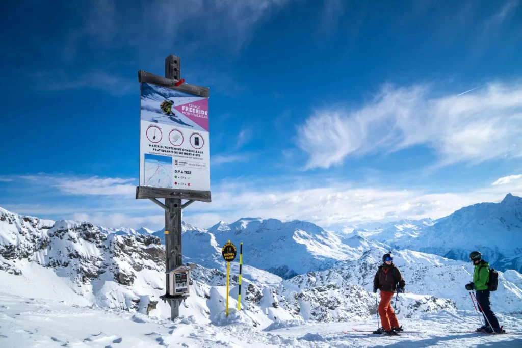 two skiers stand at the top of a ski area by a huge sign signalling a freeride zone