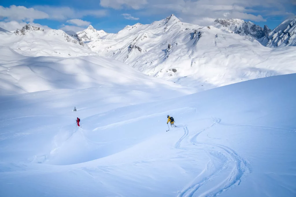 two skiers made fresh tracks at the top of a very white mountainscape, skiing in the shade