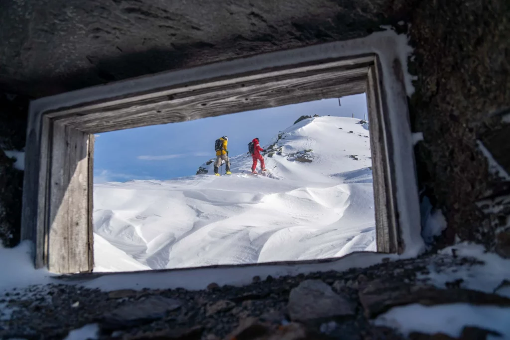 a photo of two ski tourers is taken through the small window of a small, snow-crusted mountain refuge
