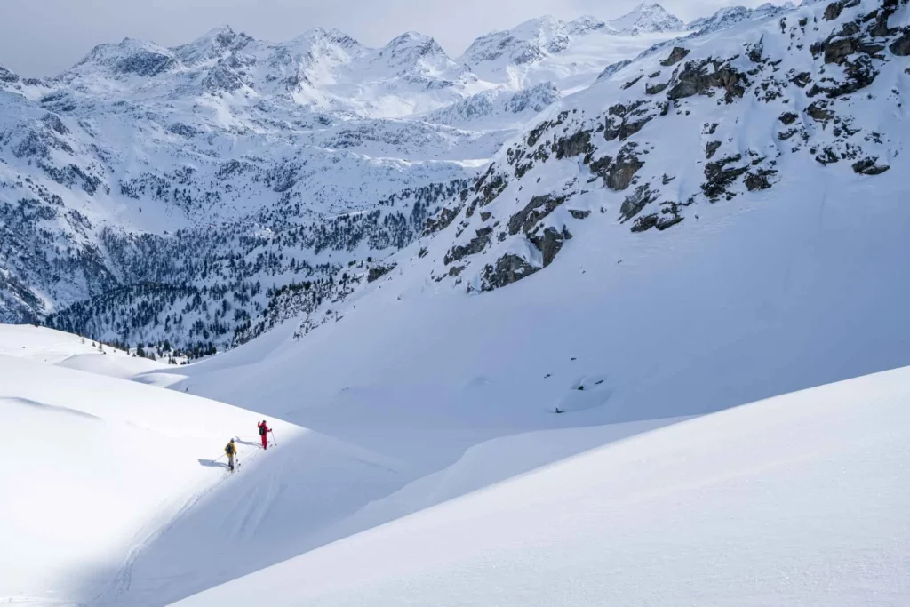two skiers look small in against a huge mountain vista in a very snowy environment