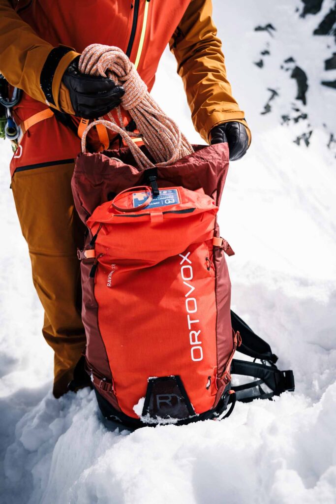 a person in red, pulls a large rope out of a red Ortovox backpack resting on snow