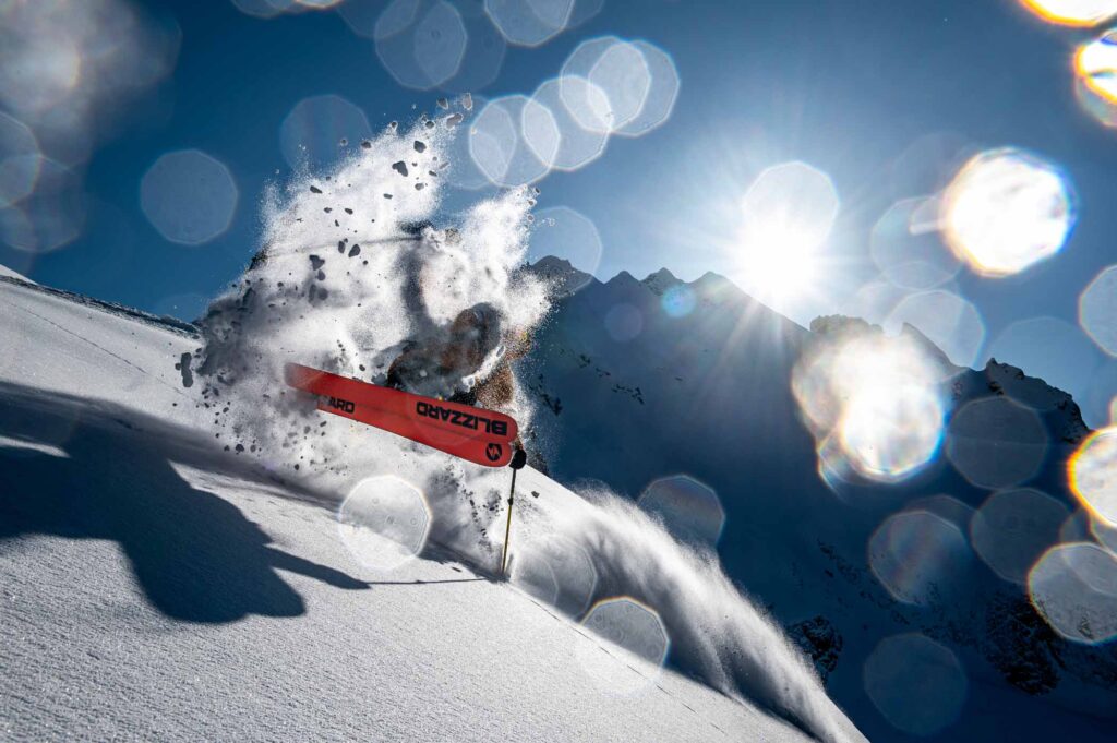 a skier takes a little air on a turn in fresh snow, hiding themselves from camera view with powdery spray, orange ski bases dominating the image