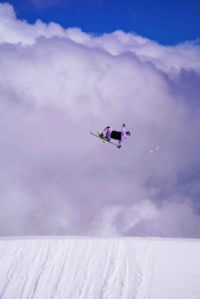 skier in purple is shot in the air, mid-grab and flip, having taken off from a kicker. The photo is particularly cool, with the skier, sideways, against the cloud behind