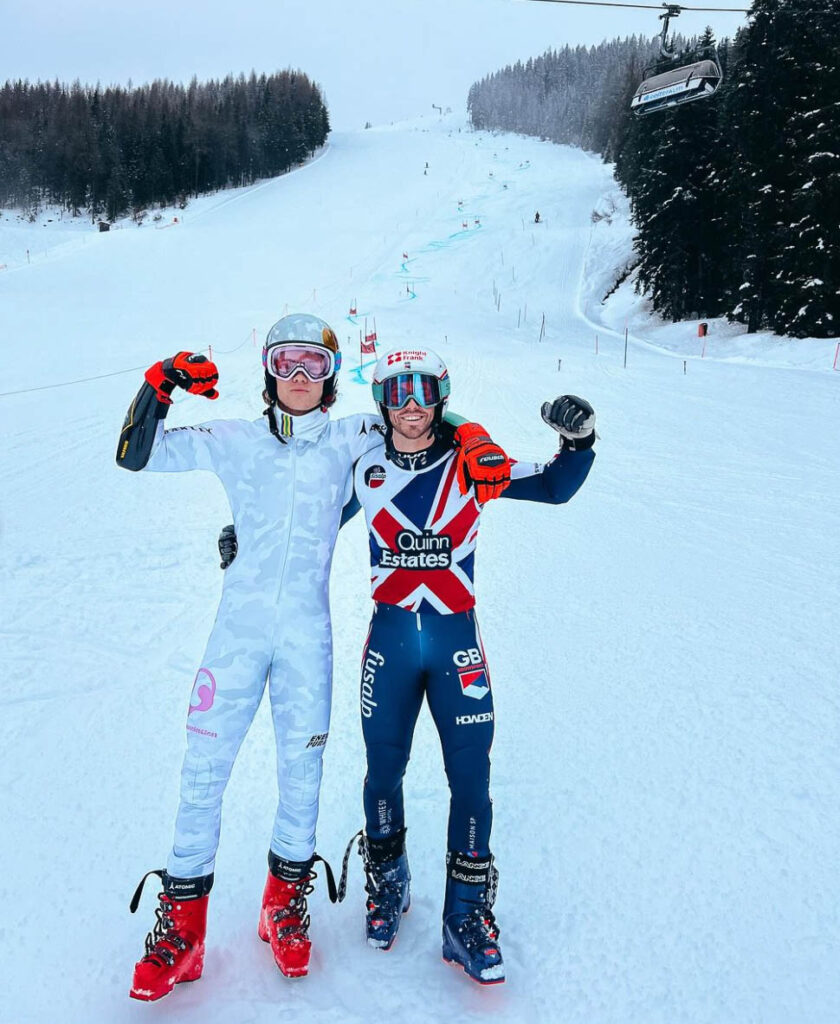 two ski racers in skins, one British Raposo and Norwegian-Brazilian Braathen, stand on slope, arms around each other, punching the air with free arm