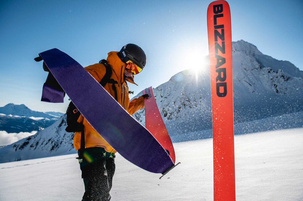 man pulls off a purple skin from an orange ski base at the top of a mountain on a blue sky day