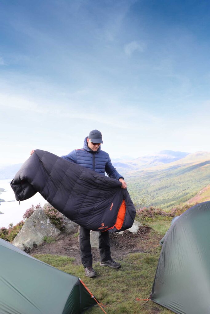 man holds sleeping bag outside tent, on a hillside